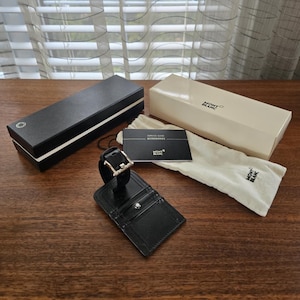 Peut inclure: Un porte-étiquette de bagage en cuir noir avec une boucle argentée et un emblème Montblanc, à côté d'une boîte noire, d'une boîte crème, d'une pochette blanche et d'un guide de service. Sur une surface en bois.