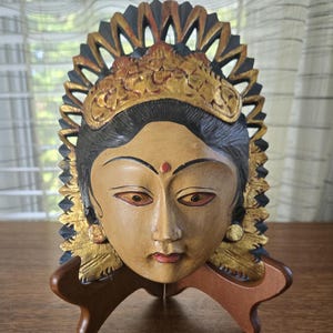 May include: A carved wooden mask featuring a serene face with tan skin, dark hair, and a red bindi. The mask is adorned with a gold-painted crown and circular earrings, set against a black, fan-shaped, openwork decorative border on a brown stand.