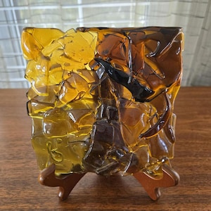 May include: A decorative glass vase with a textured, mosaic-like design in shades of amber and brown. The vase is mounted on a wooden stand, showcasing its unique, artistic appearance. The glass has a layered, irregular surface.