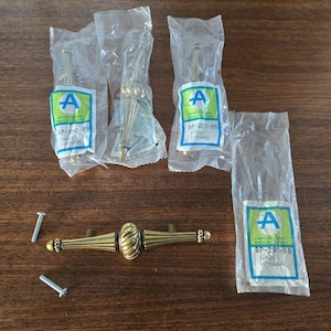May include: A collection of vintage brass cabinet pulls and knobs, some still in their original packaging. The pulls have a detailed, ornate design, and the knobs have a simple, elegant shape. The packaging includes the text "A" and "BP-290-BB".