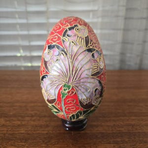 May include: An ornate, egg-shaped decorative object. The egg features a red background with gold accents and a central floral design in shades of pink and purple. The egg is mounted on a small, dark wooden stand, showcasing its intricate details.