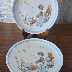Może przedstawiać: Dwa białe ceramiczne talerze z jasnoniebieską obwódką. Każdy talerz przedstawia fantazyjną ilustrację dziecka puszczającego latawiec na polu z grzybami i kwiatami. Uroczy, stylizowany na vintage design.