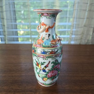 Puede incluir: Un pequeño jarrón de porcelana blanca con un diseño floral y de pájaros colorido. El jarrón tiene un cuello estrecho y una base ancha, y está decorado con una variedad de colores, incluyendo rojo, verde, azul y amarillo. El jarrón tiene un par de asas con forma de dragones.