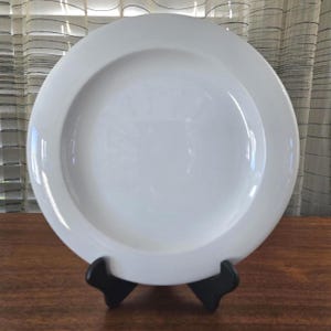 May include: A plain white circular dinner plate with a wide, smooth rim and a slightly recessed centre. The plate is held upright by a black plastic display stand, showcasing its clean, minimalist design and glossy, reflective surface finish.
