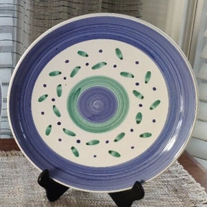 May include: A large, white ceramic plate with a blue and green design. The plate has a central circle with a swirl pattern, surrounded by a ring of green leaves and blue dots.