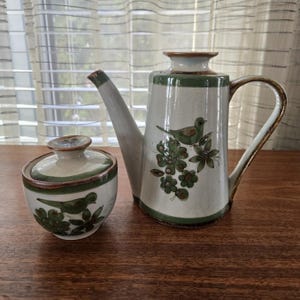 May include: A ceramic teapot and lidded sugar bowl set. The off-white pieces have green and brown trim, with a bird and floral design. The teapot has a curved handle and spout. The sugar bowl is round.