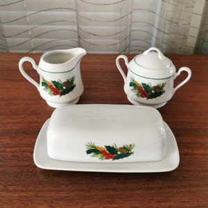 Può includere: Un set da portata in ceramica bianca con una lattiera, una zuccheriera con coperchio e una burriera. Ogni pezzo è decorato con un bordo verde e un motivo festivo di agrifoglio, bacche e pigne. Il set è esposto su una superficie di legno.
