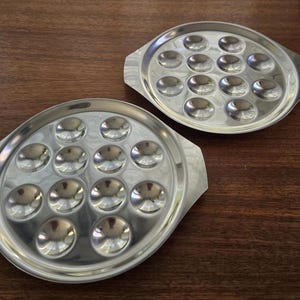 Puede incluir: Dos bandejas de servir de metal plateado con asas. Cada bandeja tiene 12 pequeñas hendiduras redondas para servir aperitivos o refrigerios.