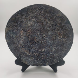 May include: A blue ceramic plate with a raised lace design. The plate is on a black stand.