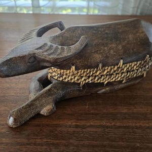 Puede incluir: Escultura de búfalo de agua de madera tallada en tonos marrones. La escultura presenta cuernos, una cara detallada y una banda tejida alrededor del cuerpo. La pieza se exhibe sobre una superficie de madera.