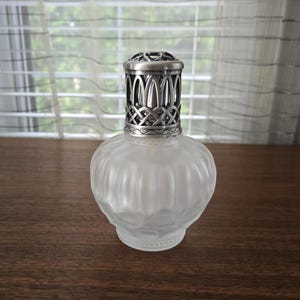 Puede incluir: Una lámpara de fragancia de vidrio esmerilado con un cuerpo redondeado y acanalado, y una tapa decorativa de metal plateado con un intrincado diseño de celosía. La lámpara tiene una base ancha y un cuello estrecho con una cubierta perforada.