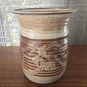 May include: A ceramic vase with a wide rim and a textured surface. The vase is in shades of beige and brown. The lower section has an embossed floral pattern. The vase is suitable for displaying flowers or as a decorative item. The height is approximately 20 cm.