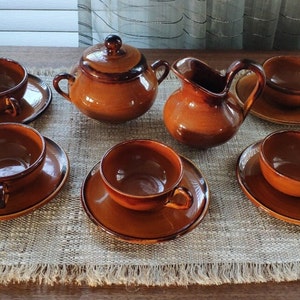 Pode incluir: Um conjunto de chá completo em cerâmica marrom, incluindo seis xícaras com pires, um açucareiro com tampa e um cremeira. O conjunto é disposto sobre um tapete de mesa tecido, mostrando uma estética rústica e artesanal.