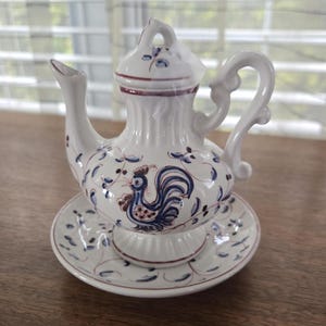 May include: White ceramic teapot with a matching saucer. The teapot features a rooster design in blue and red, with blue floral accents. The handle and spout are curved. The saucer has a matching blue floral pattern.