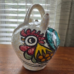 Może przedstawiać: Biały dzbanek ceramiczny z wzorem koguta. Kogut jest pomalowany na czerwono, żółto, zielono i niebiesko. Dzbanek ma dzióbek i uchwyt.