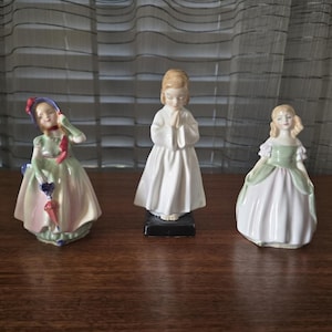 Può includere: Tre statuine di porcellana di giovani ragazze. Una ragazza tiene un ombrello e indossa un vestito verde e rosa. Un'altra indossa un vestito bianco con le mani giunte in preghiera. La terza ragazza indossa un vestito verde e bianco.
