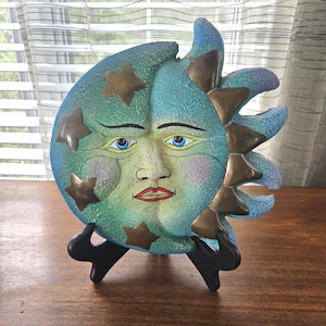 May include: A decorative wall plaque shaped like a crescent moon with a human face, featuring a textured blue and green finish, metallic gold stars, and gold sun rays on the right side, displayed on a black plastic stand for upright support.