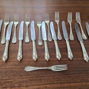 May include: A collection of vintage cutlery, including forks and knives, displayed on a wooden table. The silverware features decorative handles and polished blades. The forks have three or four prongs. The knives have a pointed end. Ideal for a formal dinner setting.