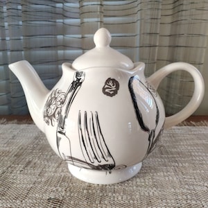 Puede incluir: Tetera de cerámica blanca con un diseño en blanco y negro de utensilios de cocina. La tetera tiene un cuerpo redondo y una tapa con un pomo en la parte superior. El diseño incluye un tenedor, un cuchillo y una cuchara.