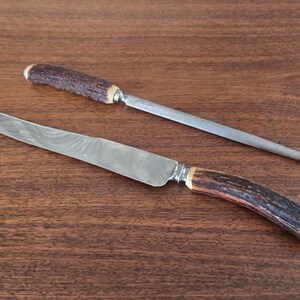 以下が含まれることがあります： 木製の表面に、彫刻刀と研ぎ棒が置かれています。ナイフは模様入りの刃と茶色の柄をしています。研ぎ棒は茶色の柄をしています。