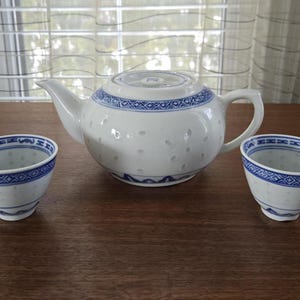 Op de afbeelding: Een witte theepot met blauwe en witte bloemenmotieven en twee bijpassende theekopjes. De theepot heeft een rond lichaam en een tuit aan de zijkant. De theekopjes zijn klein en hebben een ronde vorm.