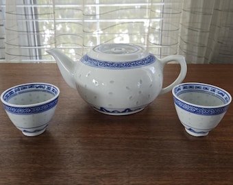 May include: A white teapot with blue and white floral designs and two matching teacups. The teapot has a round body and a spout on the side. The teacups are small and have a round shape.