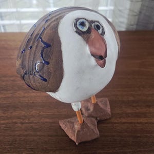 Może przedstawiać: Kapryśna ceramiczna rzeźba ptaka o zaokrąglonym korpusie, z brązowymi, białymi i niebieskimi akcentami. Ptak ma duże, wyraziste oczy i charakterystyczny dziób. Stoi na dwóch kwadratowych, brązowych stopach z pomarańczowymi nogami.