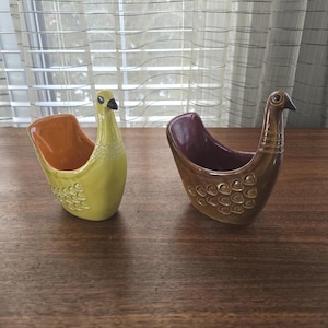 May include: Two ceramic bird-shaped planters. One is yellow with an orange interior, the other is brown with a purple interior. Both have black beaks and decorative detailing. They sit on a wooden surface.
