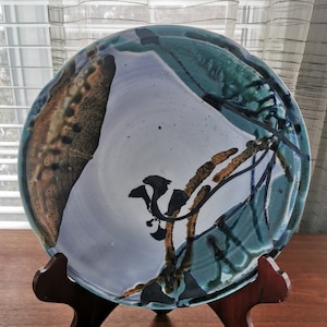 May include: A large ceramic plate with a white background and abstract blue, green, and brown designs. The plate is on a wooden stand.