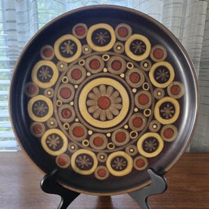 May include: A brown ceramic plate with a circular design featuring concentric circles and floral patterns in shades of yellow, brown, and red. The plate is displayed on a black stand, showcasing its decorative design.