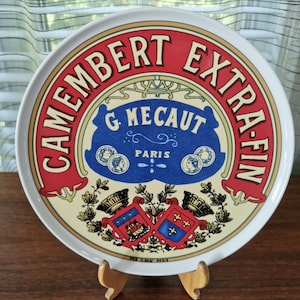 Puede incluir: Un plato redondo de cerámica con una etiqueta de estilo vintage de Camembert Extra-Fin. El diseño incluye una banda roja con texto en crema, una forma azul central con G. Mecaut Paris, dos pequeños retratos y dos escudos heráldicos.