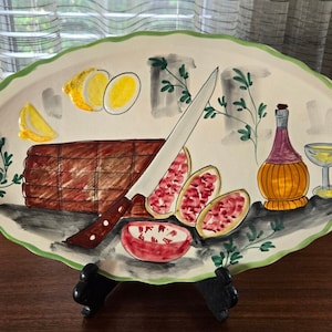 May include: A white oval ceramic serving platter with a green rim, featuring a hand-painted design of a loaf of bread, a knife, lemons, pomegranates, a bottle of wine, and a wine glass.