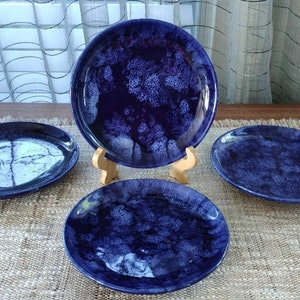 以下が含まれることがあります： 白い大理石模様の青いセラミックプレート4枚。プレートは茶色の織り生地の表面に正方形に配置されています。