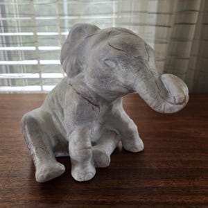 May include: A grey elephant figurine in a seated pose. The sculpture has a textured surface with visible cracks and a trunk curled downwards. The elephant's ears are large and rounded, and the overall design is detailed.