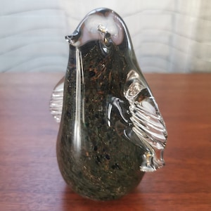 Puede incluir: Una figurita de pingüino de vidrio verde oscuro con un ala de vidrio transparente. El pingüino tiene una superficie texturizada y moteada.