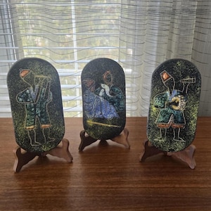 Op de afbeelding: Drie decoratieve, ovale keramische platen met geschilderde figuren, elk op een houten standaard. De platen tonen figuren in kleurrijke kleding die instrumenten bespelen. De achtergrond is donker met een gespikkelde textuur.