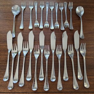 May include: A collection of stainless steel silverware, including forks, knives, and spoons, arranged on a dark wooden surface. The silverware has a simple, elegant design, reflecting light. The set includes various sizes of forks and knives.