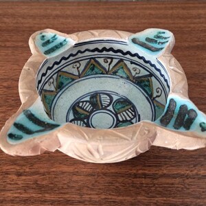 May include: A decorative, hand-carved ashtray with a unique design. The ashtray features a central bowl with a painted, intricate pattern in teal, gold, and black. The exterior is a light beige color with four rounded extensions.