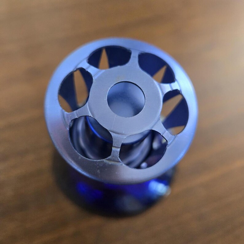 Pu&ograve; includere: Primo piano di un oggetto metallico blu dal design circolare. L'oggetto presenta forme ritagliate nella parte superiore e un foro centrale. Si trova su una superficie di legno. Diametro di circa 2,5 cm.