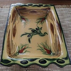 Pode incluir: Um prato de cozedura retangular de cerâmica com um esmalte amarelo e desenhos pintados de ramos de oliveira verdes e castanhos. O prato tem uma borda ondulada e uma borda festoneada.