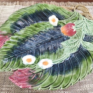 Può includere: Un piatto da portata in ceramica verde e rosso a forma di foglia di fragola con fiori bianchi e una fragola rossa.