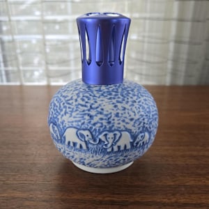 May include: A blue and white ceramic fragrance lamp with a decorative elephant design. The round body has a textured pattern and a row of elephants. The top is a metallic blue cap with vertical slits.