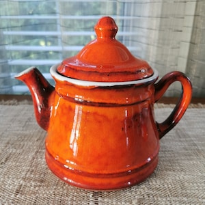 May include: A vintage orange ceramic teapot with a white rim and lid. The teapot has a rounded body and a spout on one side and a handle on the other.