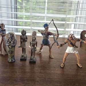 May include: A collection of six miniature figurines depicting ancient Egyptian figures, including a woman holding a cat, a pharaoh, a deity, a kneeling figure, an archer with a bow, and a warrior holding a wooden shield and a small club.