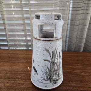 May include: A speckled white ceramic vase with a rectangular opening and a cylindrical shape. The vase features a hand-painted design of a heron and reeds in shades of grey and black. The vase is on a wooden surface.