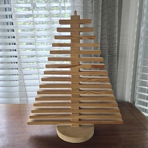 Könnte beinhalten: Eine hellbraune Holzbaumskulptur mit einem zentralen vertikalen Pfosten und horizontalen Lamellen unterschiedlicher Länge, die zu einer dreieckigen Form gestapelt sind und auf einem runden Holzsockel stehen. Die Holzmaserung ist durchgehend sichtbar.