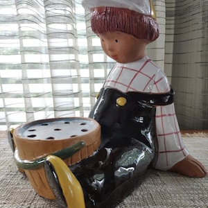 May include: A ceramic figurine of a child wearing a yellow and white hat, red and white plaid shirt, and black overalls. The child is holding a brown planter with holes in the bottom.