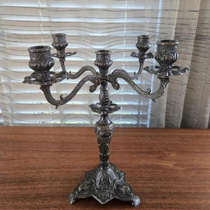 May include: A five-arm silver-toned metal candelabra featuring an ornate, textured finish with intricate scrollwork on the arms and a decorative, wide-based pedestal stand. The structure includes five candle holders arranged in a symmetrical pattern.