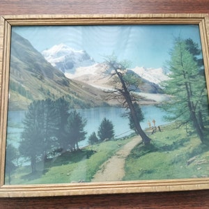 May include: A framed landscape painting depicting a mountain scene with a lake in the foreground. The painting features a snow-capped mountain in the background, a lake with a small island, and trees on the shore. The painting is framed in a gold-colored frame.