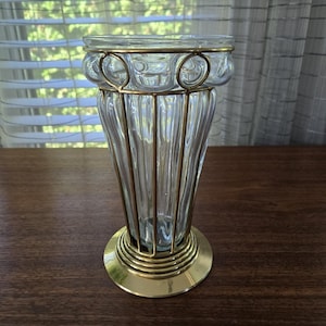 May include: A clear glass vase with a gold metal cage design. The vase has a fluted body and a round base. The top of the vase has a decorative gold metal rim. The vase is sitting on a dark brown wooden surface.